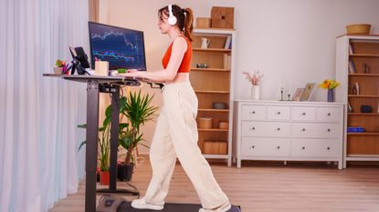 A woman walks on a walking pad at a standing desk. Behind her there are is a home setting with bookcases and plants. On her desk is a computer with graphs visible. She wears comfortable clothes, and headphones.
