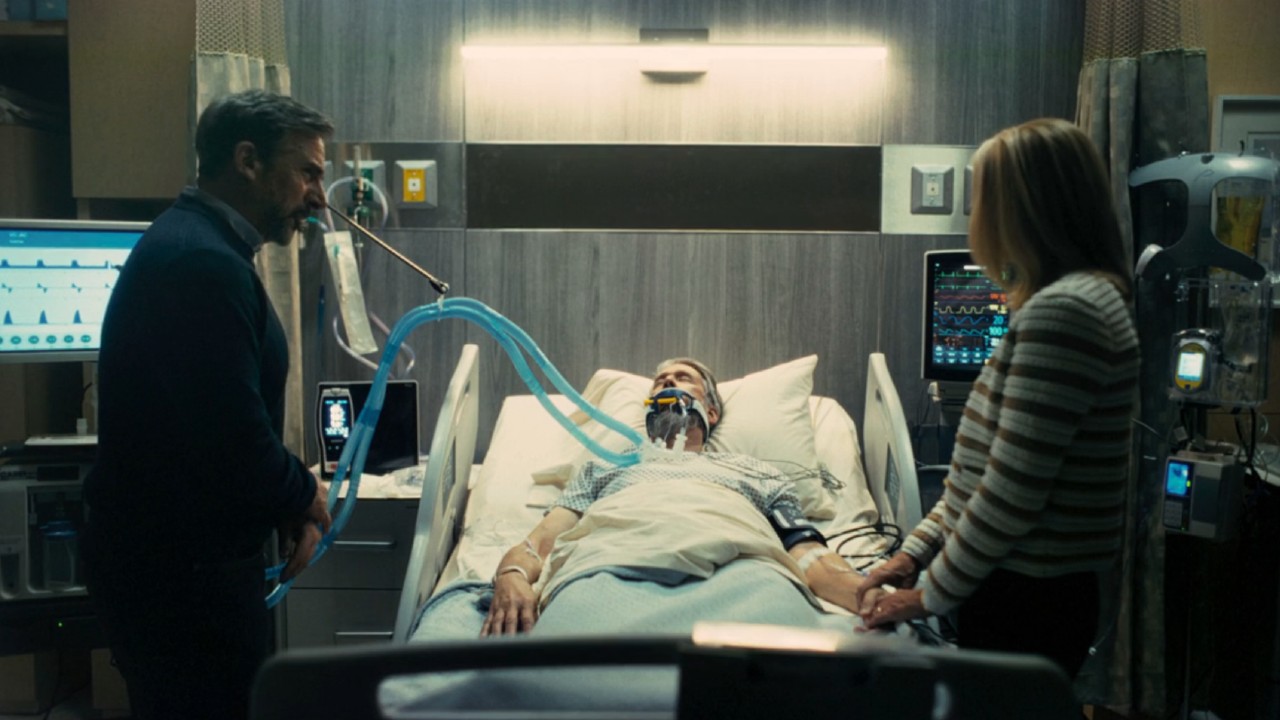 From left to right: Steve Carell standing next to a hospital bed, Alan Ruck laying in said bed with tubes down his throat, and a woman standing on the other side of the bed.