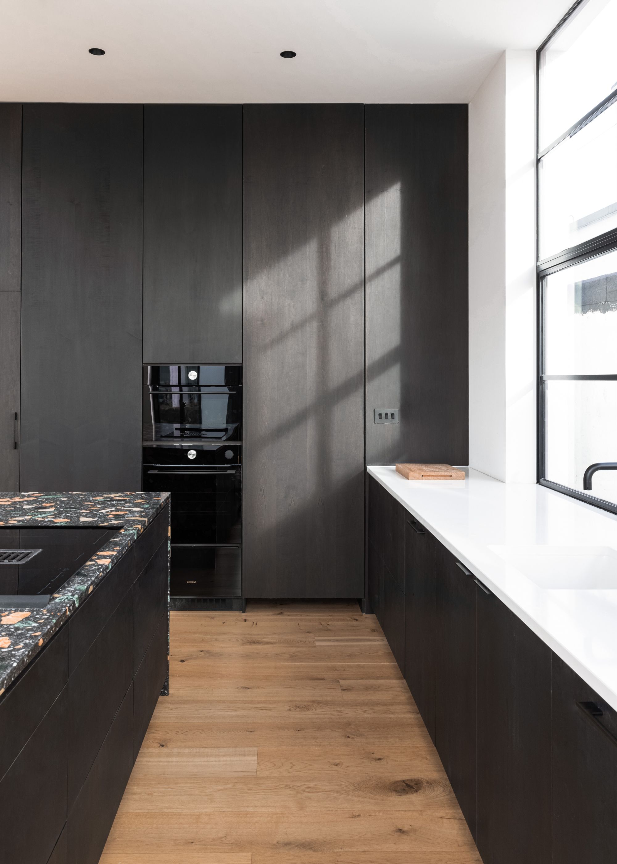 A dark small kitchen with brighten countertops and bright while painted ceilings as well as a large window to let natural light in. In this kitchen, light wooded flooring has also been used to add contrast to the space.
