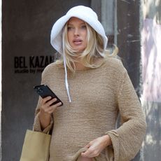Elsa Hosk wearing a white bucket hat, tan summer sweater, white bermuda shorts, flip-flops, and a The Row Cecily Mini Tote Bag