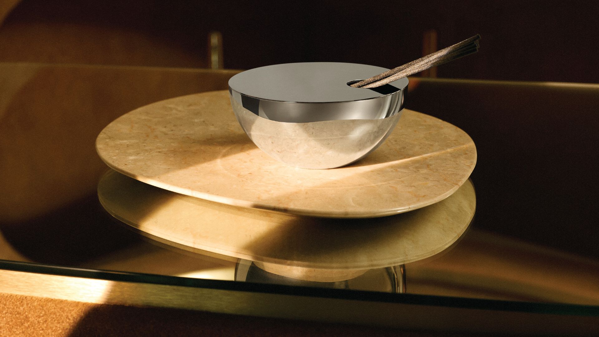 A chrome bowl reed diffuser on a beige marble trivet on a glass table