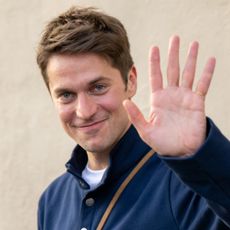 Emily in Paris star Lucas Bravo waving while wearing a navy jacket and carrying a brown leather bag