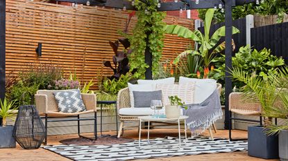 Wooden decking area backed by horizontal wood panelling and tropical plants, with wicker garden sofa and small white coffee table sat on a black and white rug