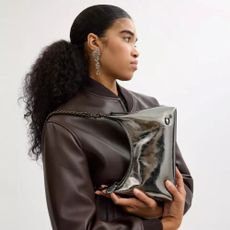 Model wearing a chocolate brown leather bomber and holding a Coach metallic handbag horizontally whilst on her shoulder