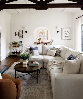 White living room with dark beams, large white sectional sofa, woven rug and wooden coffee table with plant