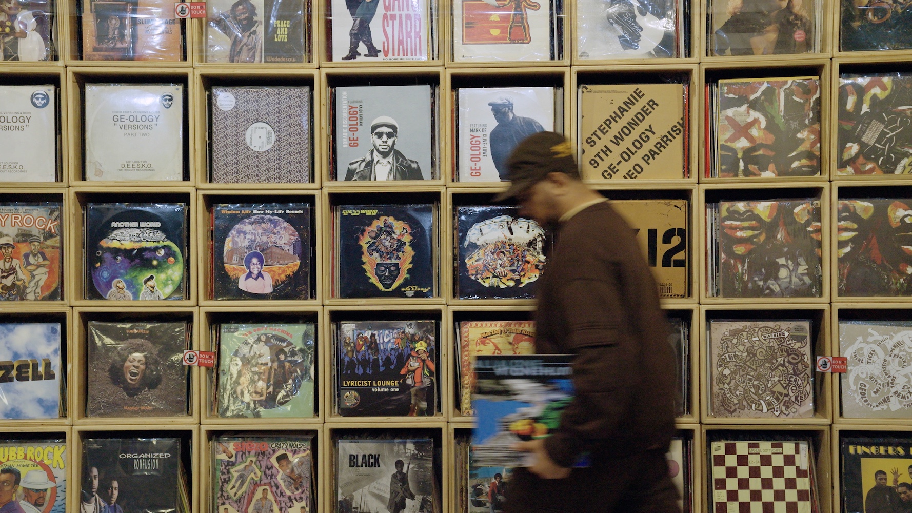 A blurred image of a man in a baseball cap walking past shelves of vinyl records with their covers showing.