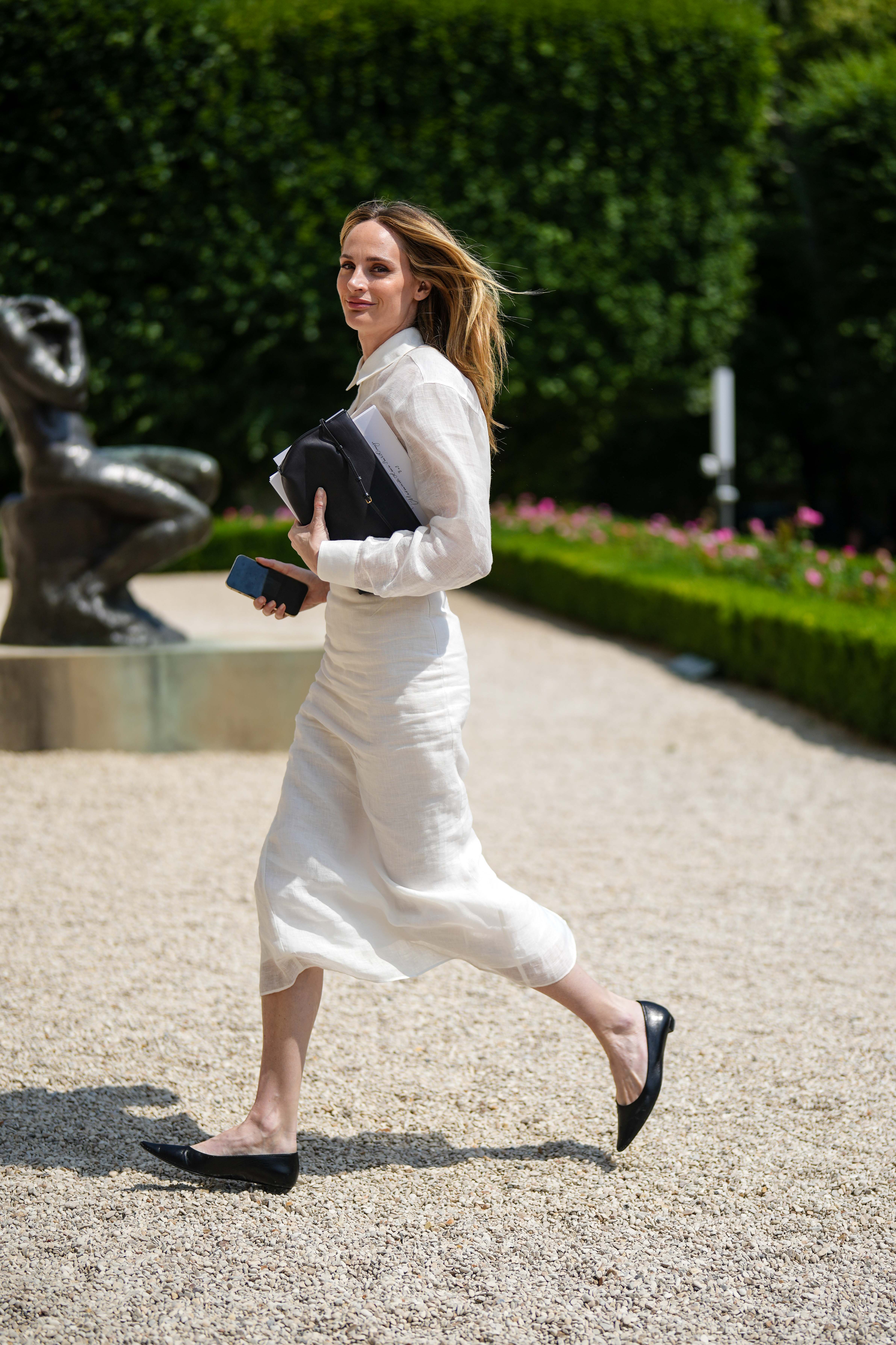 A woman wears a white linen shirt, a white linen matching midi skirt and black leather pointed ballerinas