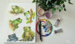 En akvarellmålning på grodor ligger på ett vitt bord bredvid lite konstnärsmaterial.