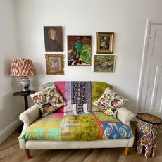 White living room with a small sofa covered with a colourful throw and vintage floral cushions, and a small gallery of painted artwork on the wall behind