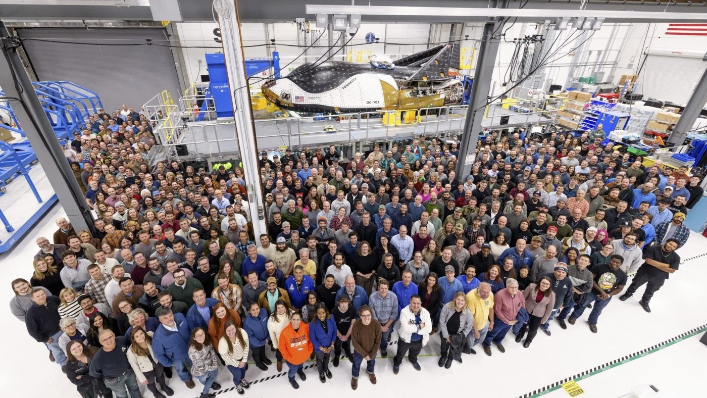 a large group of people stands in front of a small white and black space plane inside a large building.