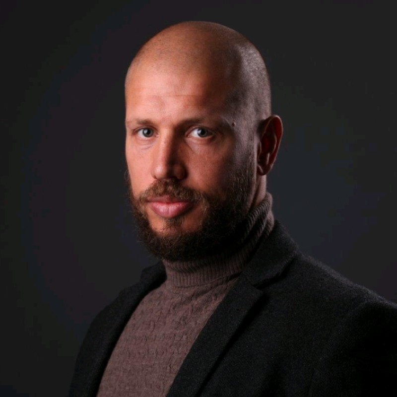 Headshot of Kelvin Elliott wearing smart black jacket and brown turtleneck against black background.