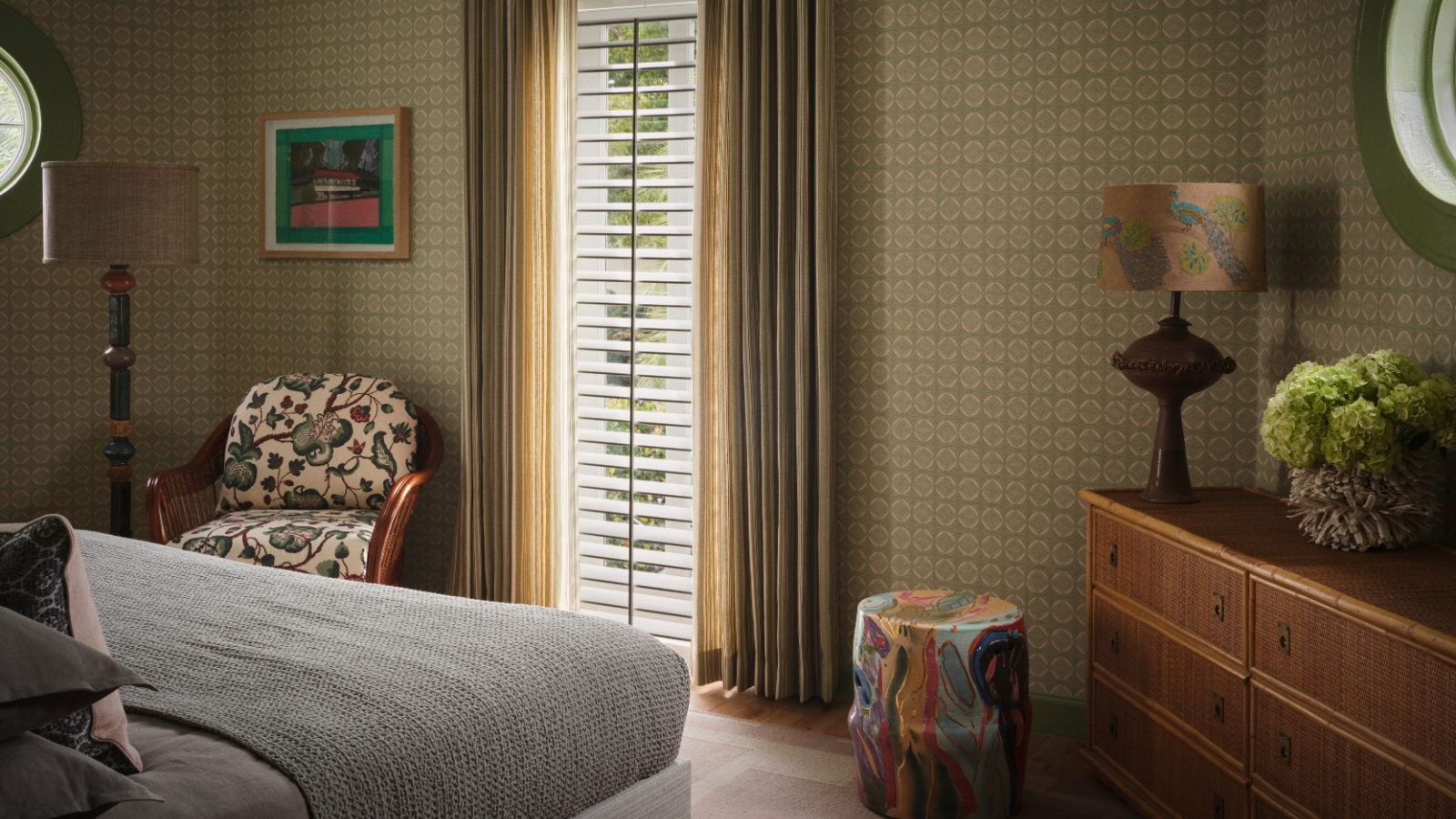 Primary bedroom with green patterned walls, woven dresser, floral armchair, colorful ceramic stool and soft layered textiles.
