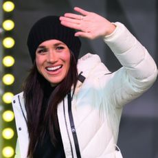 Meghan Markle waving in a white puffer jacket on stage with lights behind her 