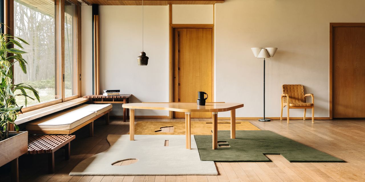 Image of a modern living room with a natural aesthetic. There are large windows on the left wall and two wooden doors on the back wall which is painted an off-white color. A bench seat is below the window and there is an abstract-shaped wooden dining table in front of it. On the floor below the table are three cutout rugs (white, green, and yellow) that fit together in a puzzle piece.