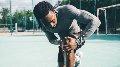 A man in running clothes kneels on a sports ground and looks closely at his knee, cupping it with two hands.