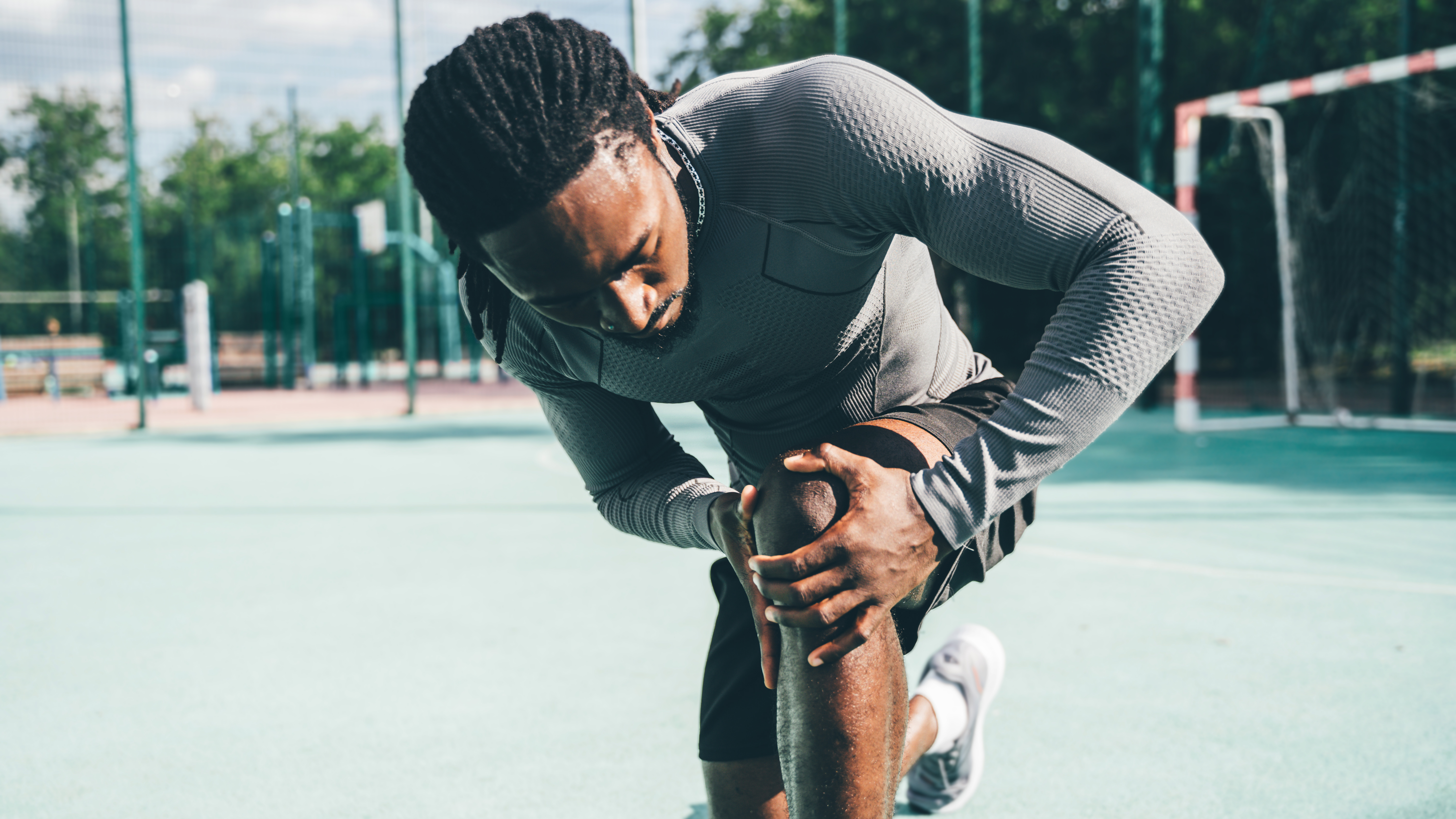 A man in running clothes kneels on a sports ground and looks closely at his knee, cupping it with two hands.