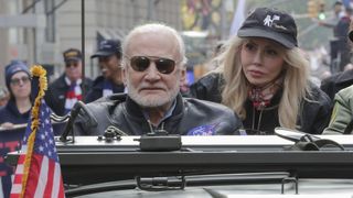 Buzz Aldrin wearing sunglasses next to Anca Faur, wearing a hat with an image of an astronaut and a flag on the front. Both are sitting in an open-top car with a crowd in behind