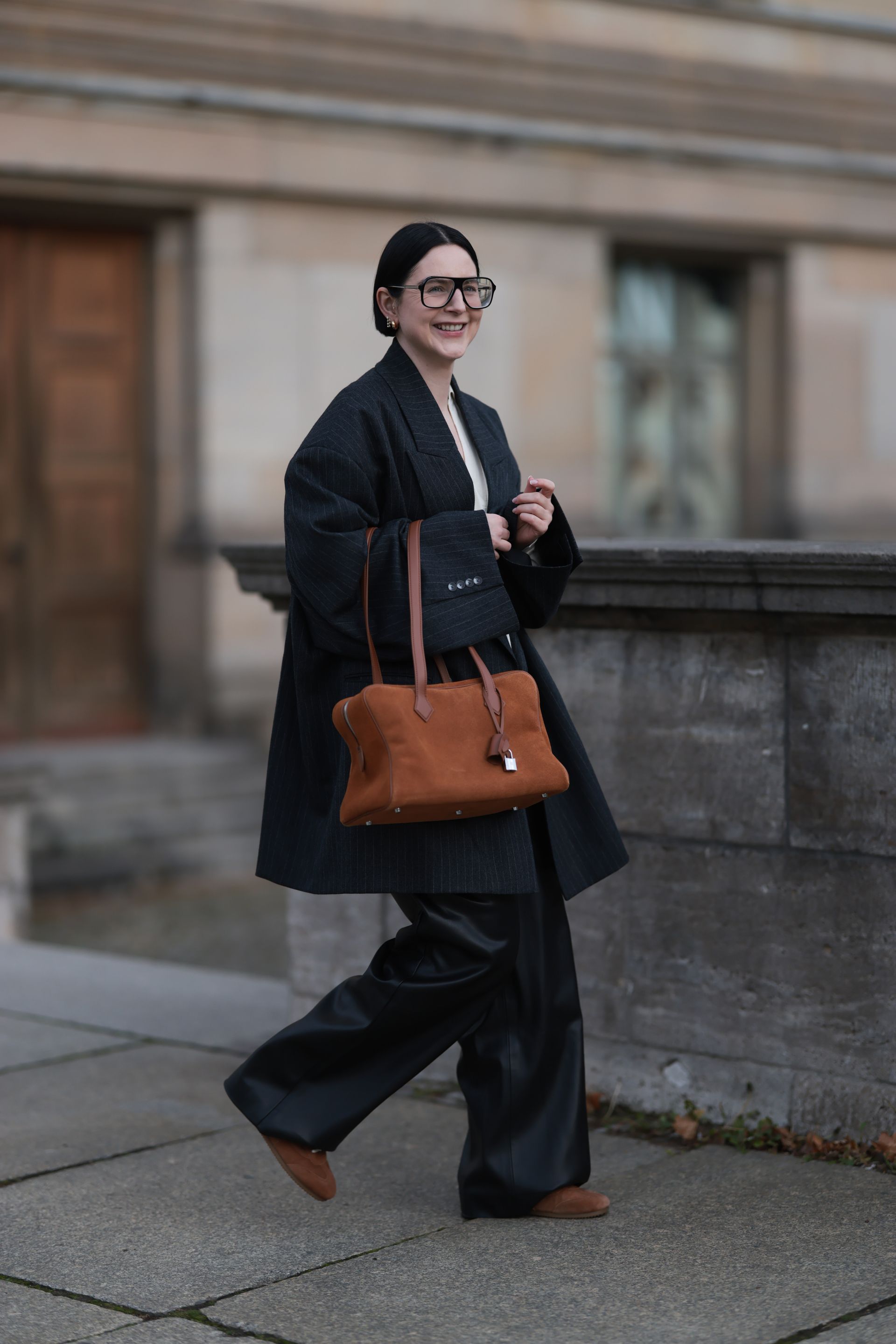 A woman wears an oversize black blazer, suede bag, black pants, and brown suede sneakers—one of the biggest sneaker color trends of 2026..