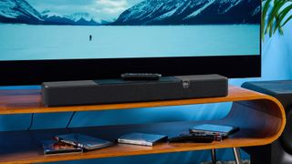 the klipsch flexus core 100 soundbar photographed on a wooden tv stand with a tv behind, showing the remote, buttons, and inputs