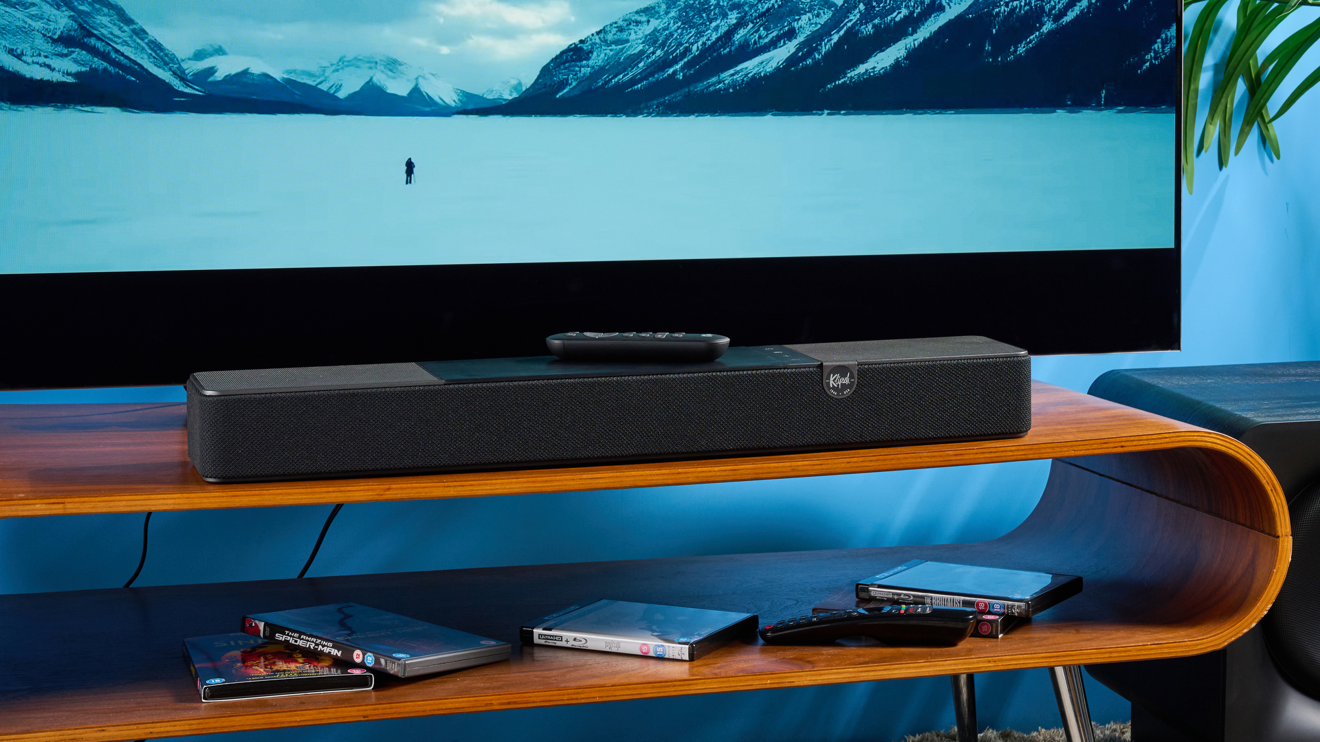 the klipsch flexus core 100 soundbar photographed on a wooden tv stand with a tv behind, showing the remote, buttons, and inputs
