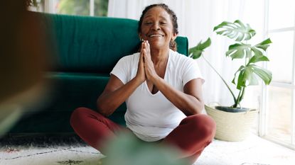 An older woman looks happy as she meditates while sitting on her living room floor.