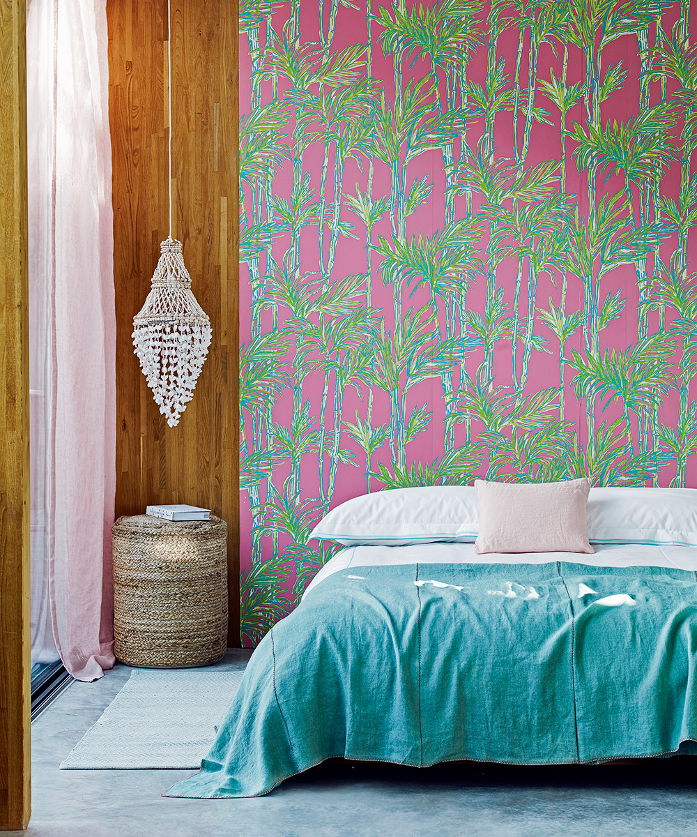 Pink and green tropical print wallpaper on a bedroom accent wall behind a white and blue dressed bed, with a shell pendant light hanging to the side.