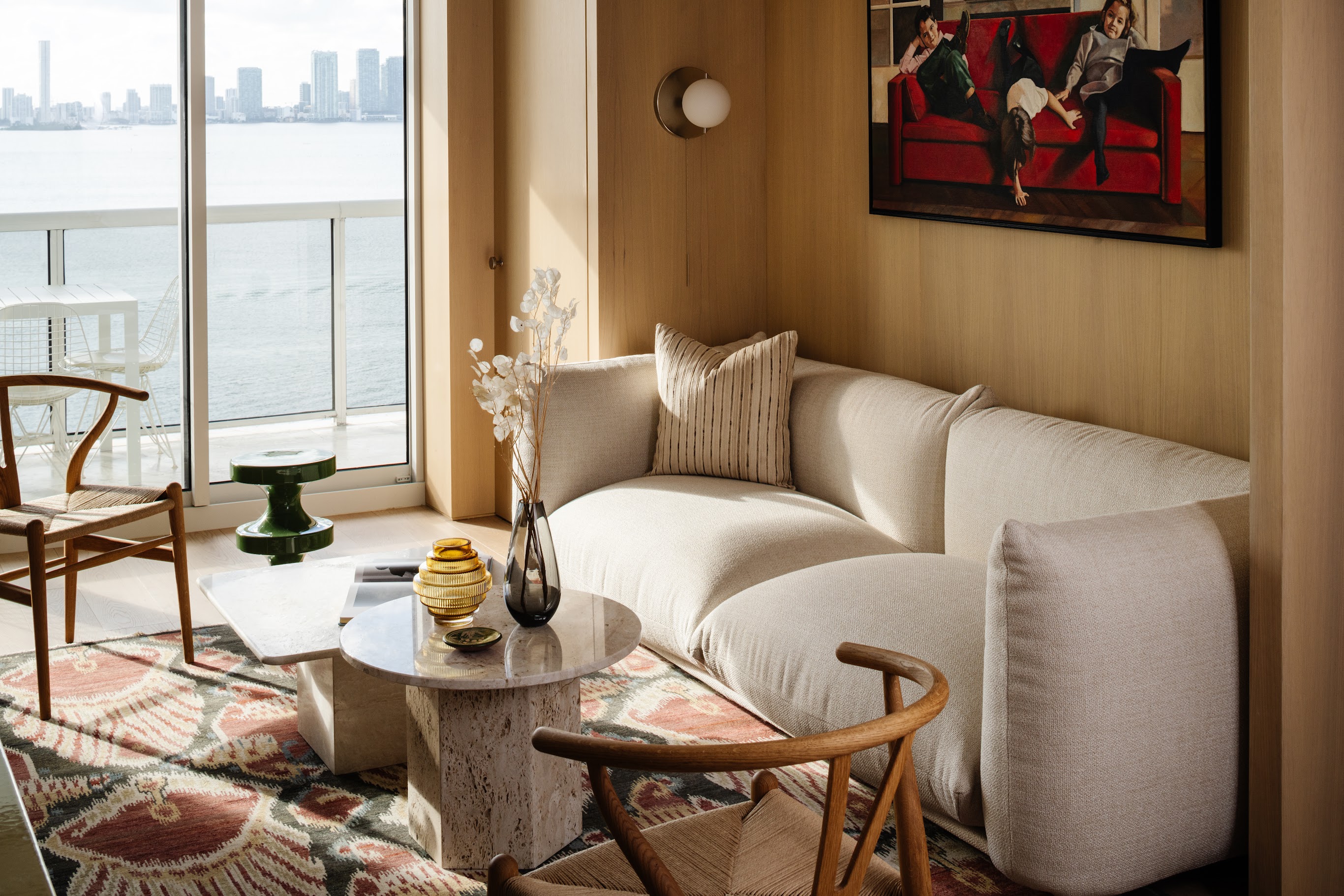 A naturally lit interior with a lounge sofa in a soft shape, wood-paneled walls, wood furniture along with marble accents, eccentric artwork, and a pendant paper lantern.
