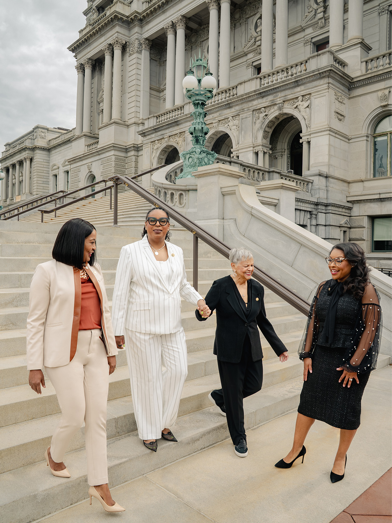 Four congresswomen walk down the Library of Congress steps together, smiling. From left: Jahana Hayes, LaMonica McIver, Bonnie Watson Coleman, and Emilia Sykes. McIver and Watson Coleman hold hands.