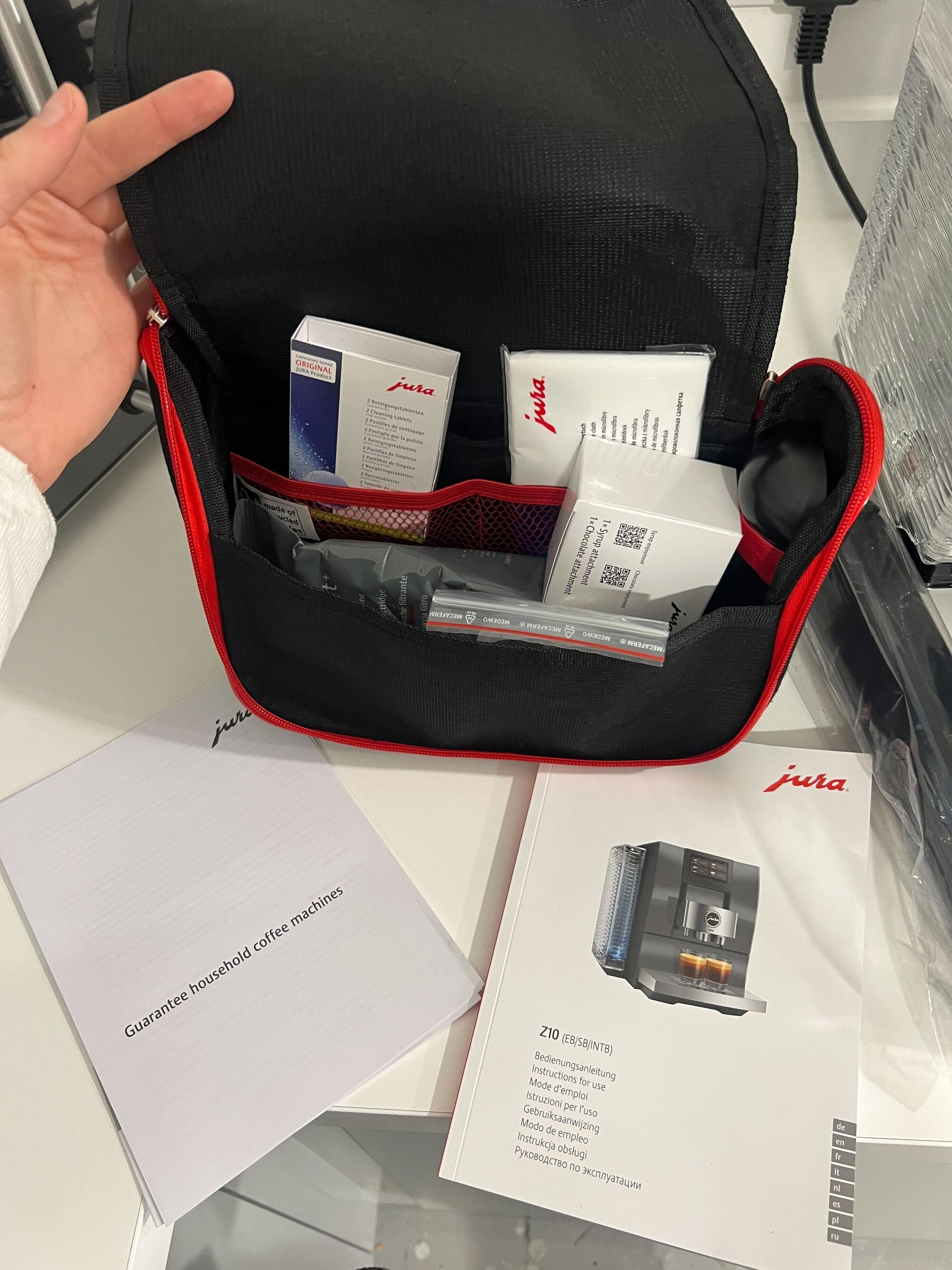 Image of a hand holding a small black and red black open that is full of white boxes that are coffee machine cleaning supplies. There are two paper direction manuals on the countertop.