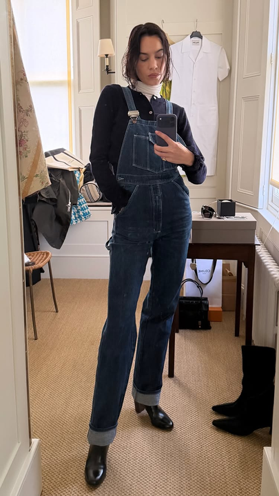 Alexe Chung takes a mirror selfie wearing layered knitwear with dungarees and rounded-toe heeled boots.