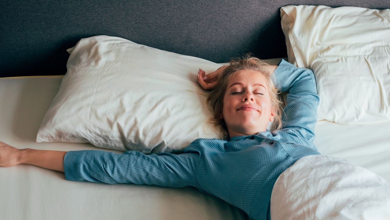 Woman lying in bed smiling with her eyes closed, with one arm behind her head and the other stretched out to the side. She is wearing blue PJs and the bedding is white.