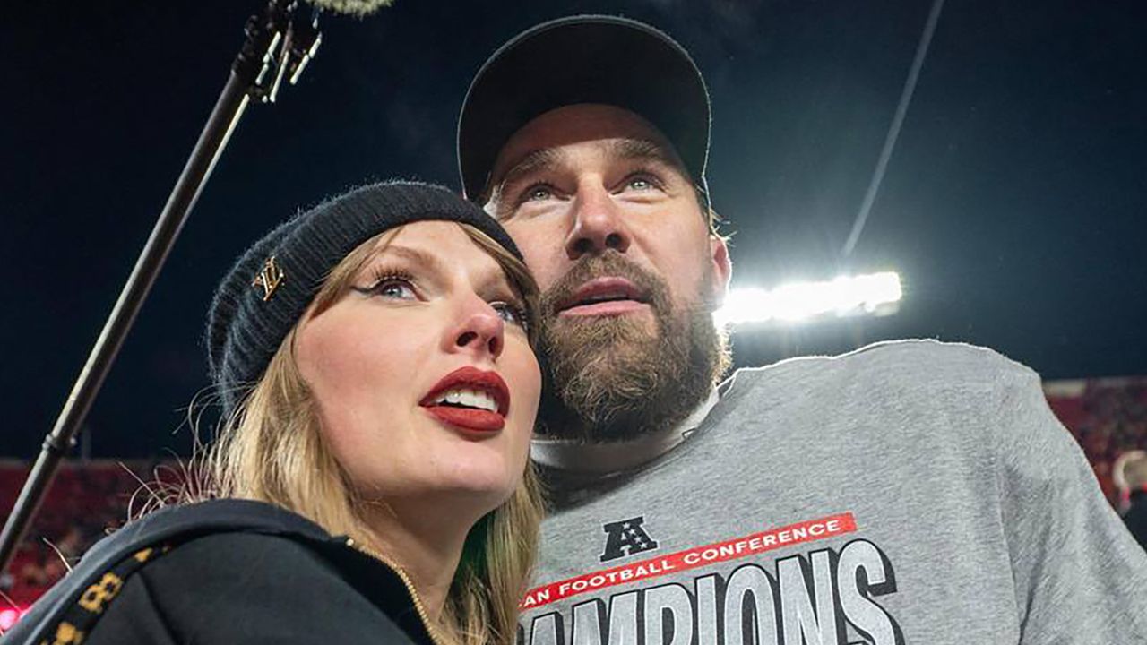 Taylor Swift wearing a black winter hat and hoodie hugging Travice Kelce on a football field with a light behind them