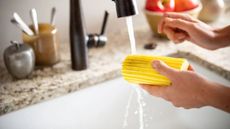 Someone rinsing a yellow Damp Duster sponge under a running black faucet into a white sink