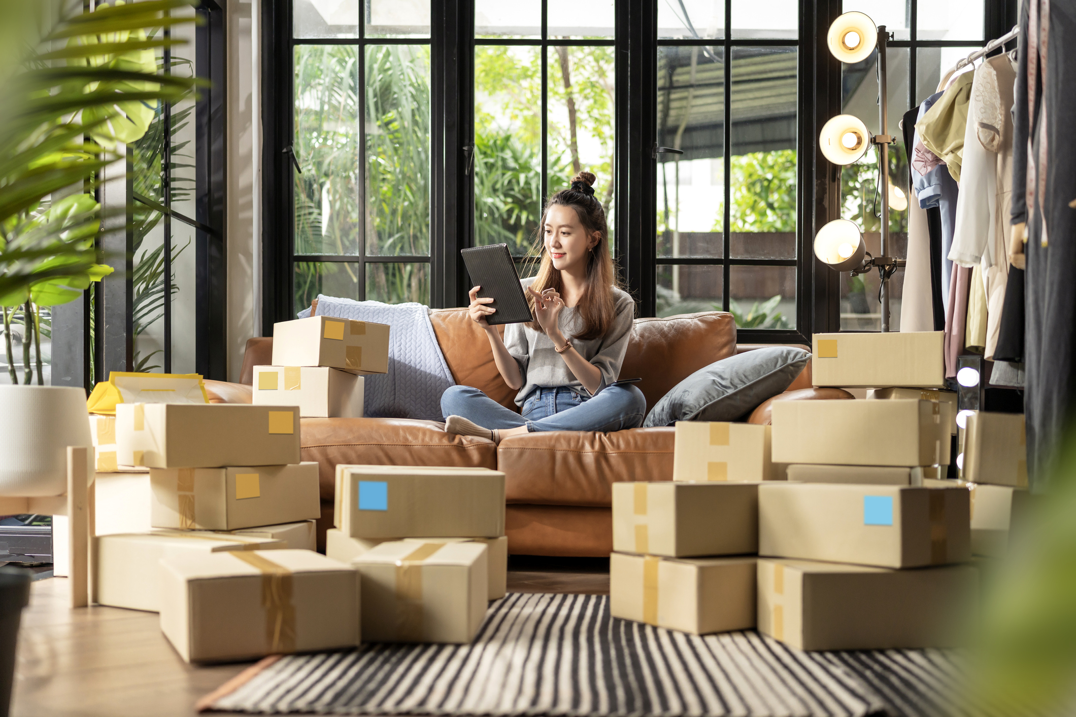 A woman sat on a sofa shopping on a tablet, surrounded by boxes from online shopping orders