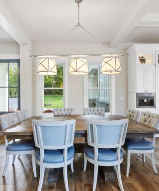 A white dining room with a three head chandelier, a dark brown wooden dining tale with eight gray and light blue chairs around the sides, two windows and a blue door, and wooden flooring