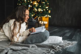 A woman sat amidst a cosy, Christmas decorated scene, while reading a book.