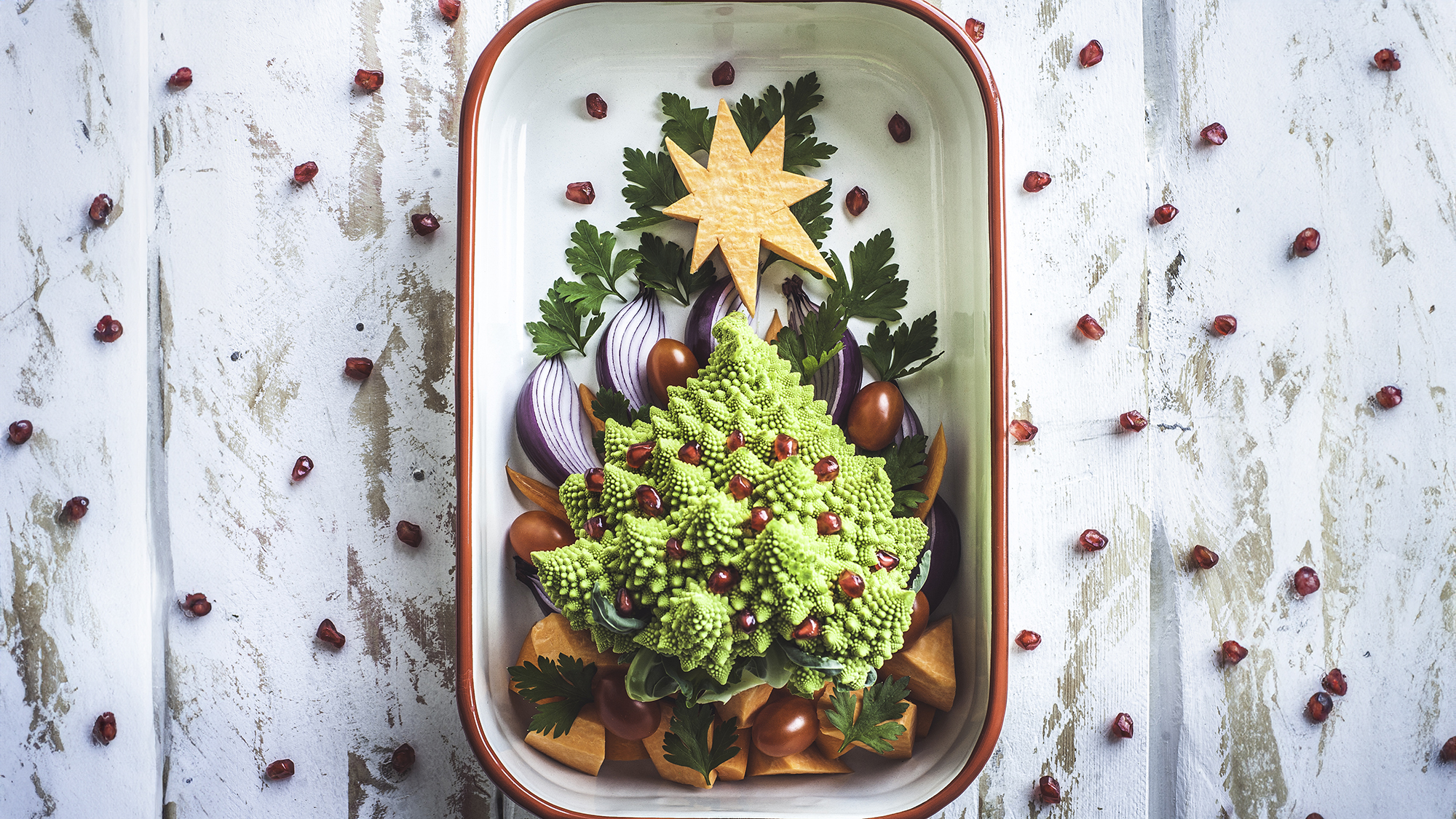 A baking tray decorated with a plant-based Christmas tree, made of Romanesco cauliflower and pomegranate arils, cherry tomatoes, red onion, sweet potato and carrots and garnished with Italian parsley. A white wooden, rustic background with a natural pattern of pomegranate seeds.