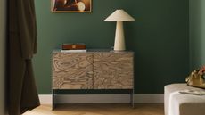 Image of a sideboard with black and white wood-grain doors and a brushed chrome frame in a dark green room. It's styled with coffee table books, lamps, and vases.
