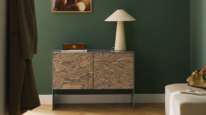 Image of a sideboard with black and white wood-grain doors and a brushed chrome frame in a dark green room. It's styled with coffee table books, lamps, and vases.