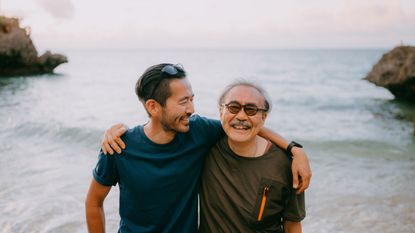 A father and his adult son smile on a beach.