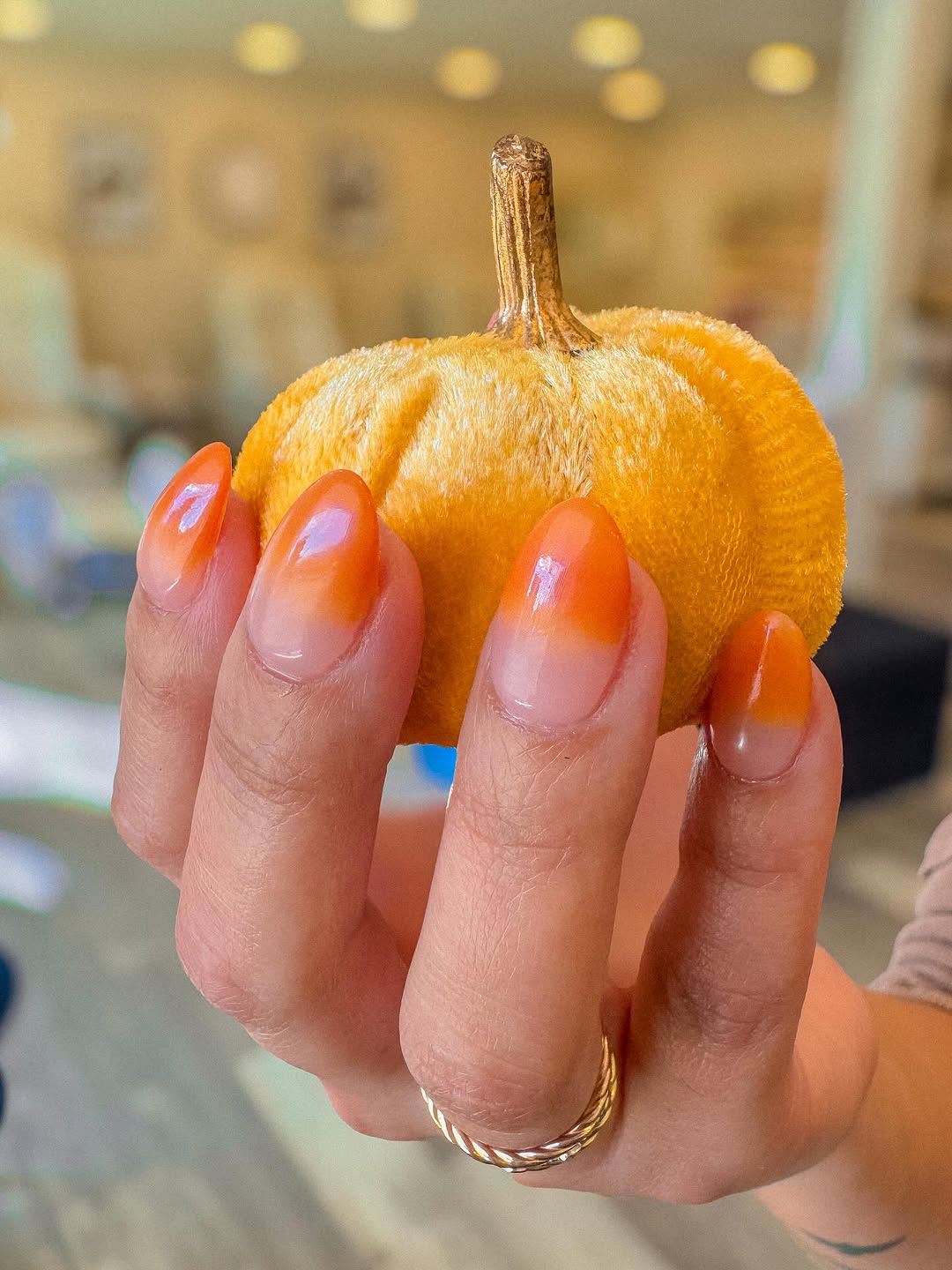 Close-up photo of a person&amp;rsquo;s hands showcasing a small pumpkin and a glossy orange ombr&amp;eacute; almond nail manicure.