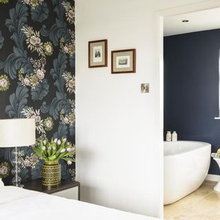 bedroom with green, black and floral wallpaper leading into an open ensuite with a freestanding white curved bathtub