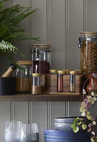 wooden spice rack with spices on wooden shelf with jars of grains, pasta, plant, pestle and mortar, bowls, glasses