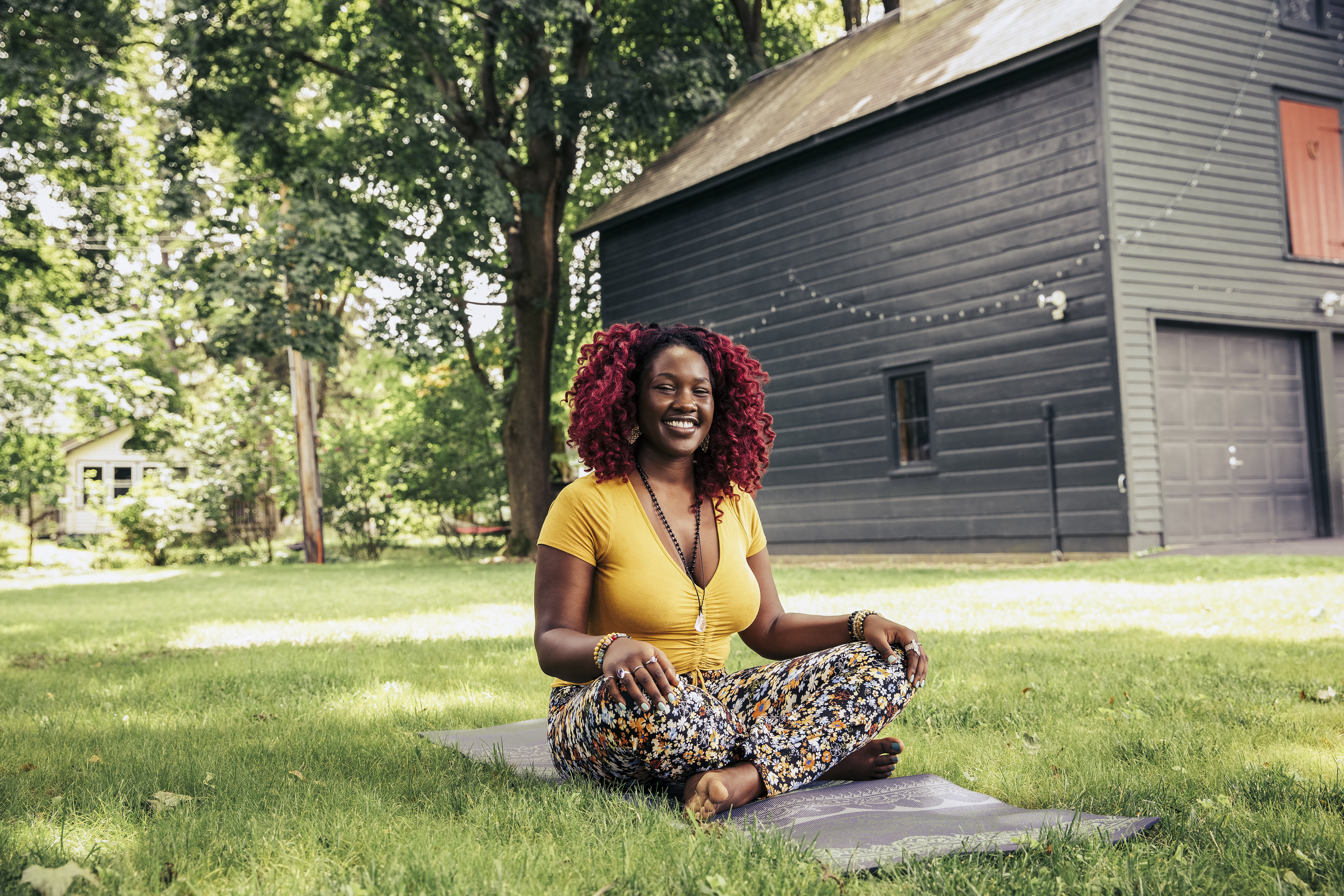 A woman sits cross-legged outside on a yoga mat. Behind her we see a large expanse of healthy-looking grass and the side of a house with a garage, which is decorated with fairy lights.
