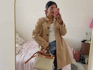 Chiara wears white top and blue jeans with tan trench coat, flats, and a handbag.