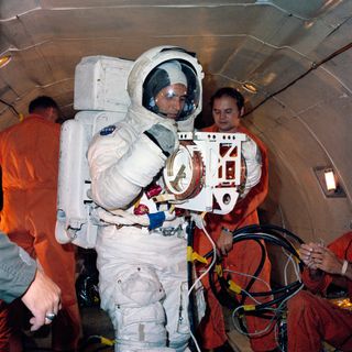 Harrison H. Schmitt during EVA training on an aircraft. He is wearing a spacesuit and people in the background are wearing orange jumpsuits.