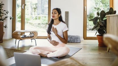 Woman sits cross-legged on a yoga mat with her eyes closed, one hand on her thorax, the other hand on her stomach