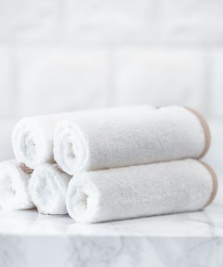 A pile of five stacked rolled white towels on a marble surface with white subway tiles and a plant behind them