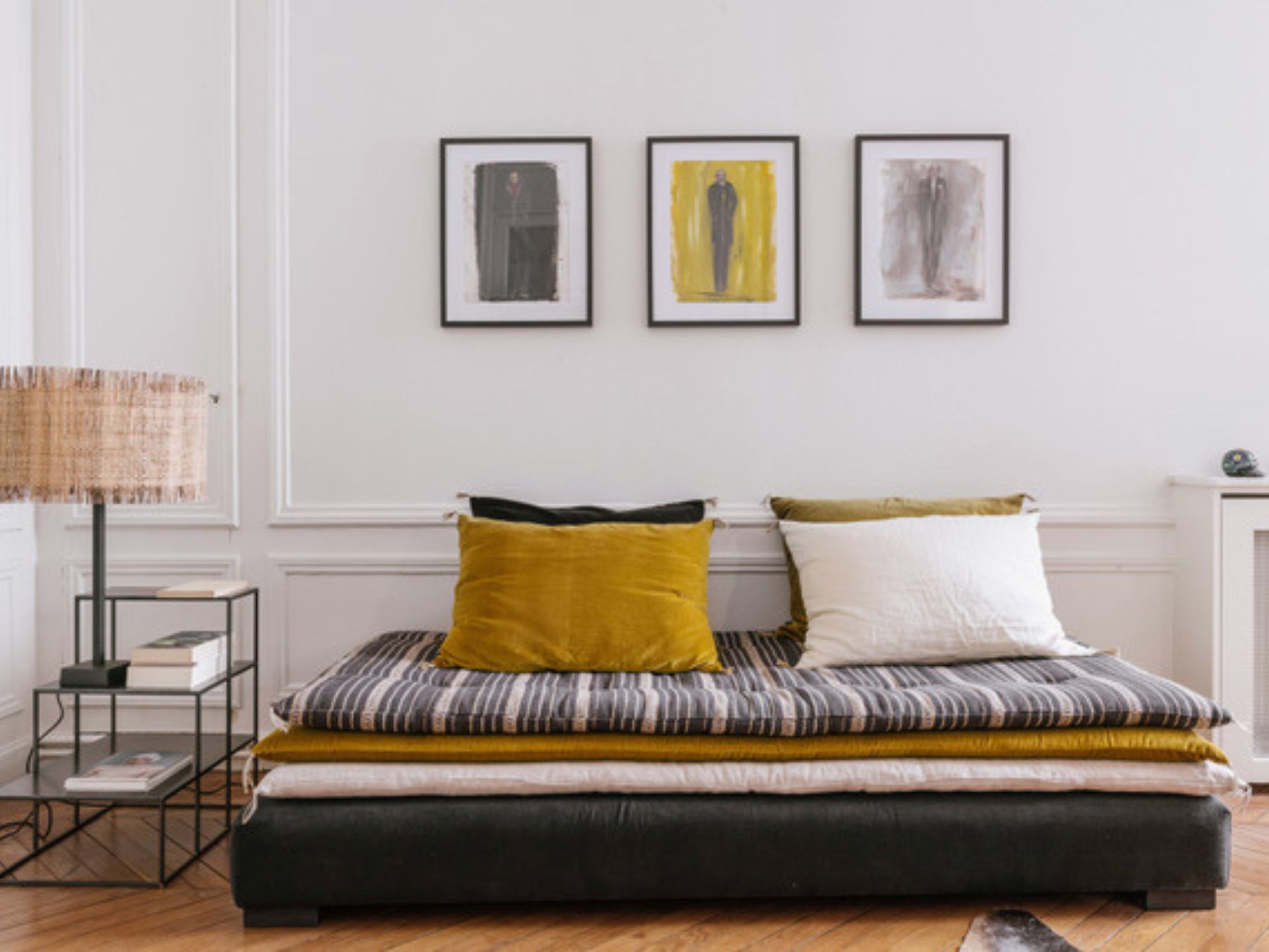 Modern white office space with wooden floors, complete with a sofa bed in white, navy and yellow/mustard cushions with a lamp and side table, panelling and three picture frames hanging on the wall