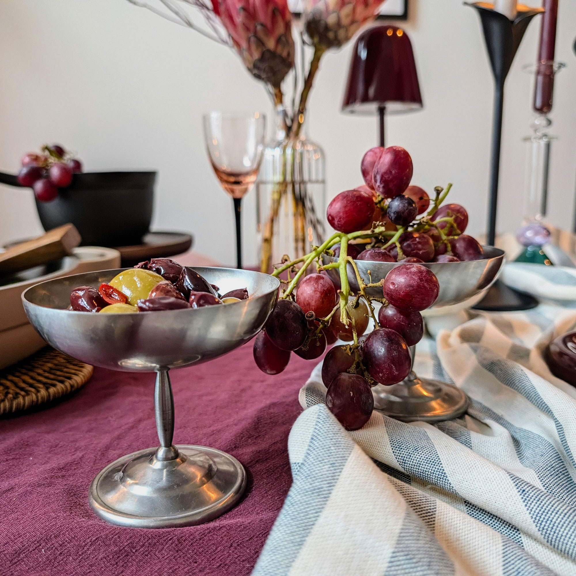 Silver coupes filled with olives and grapes on table dressed with burgundy linens and blue and white striped runner